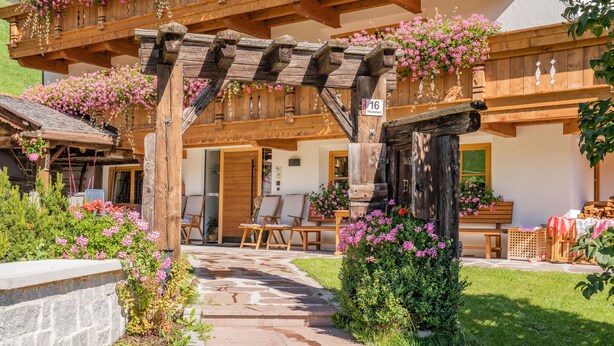 Een houten pergola met bloemenmanden, een pad van stenen en een landelijk gebouw met een houten deur en ramen.