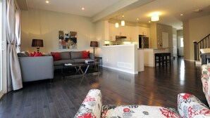 A spacious living room with a grey sofa, a coffee table, and floral-patterned chairs. A kitchen area with a central island and bar stools is visible in the background. - Boardwalk Homes - Executive Guest Rooms & Townhomes (Kitchener)