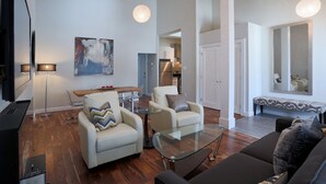 A modern living room with a sofa, armchairs, a glass coffee table, and a TV. There is a dining area with a table and chairs, and a kitchen in the background. - Boardwalk Homes - Executive Guest Rooms & Townhomes (Kitchener)
