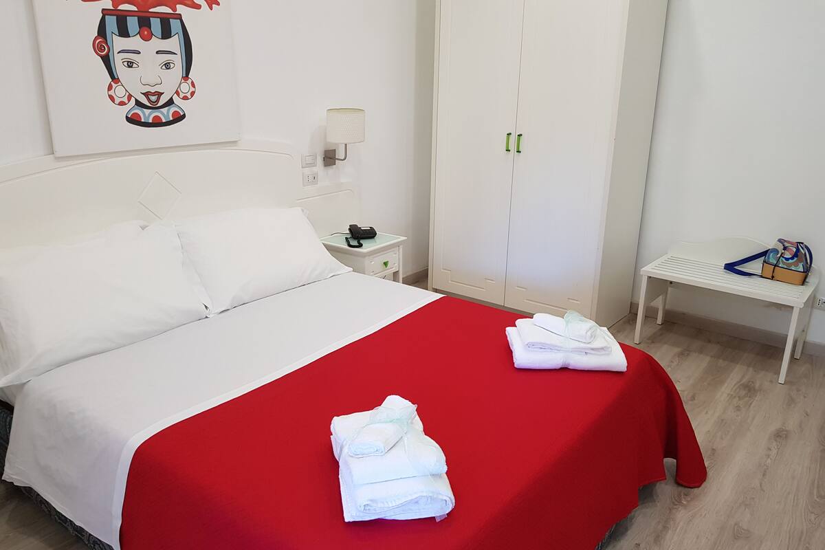 A hotel room with a bed, a bedside table with a lamp, a wardrobe, and a bench with a bag.