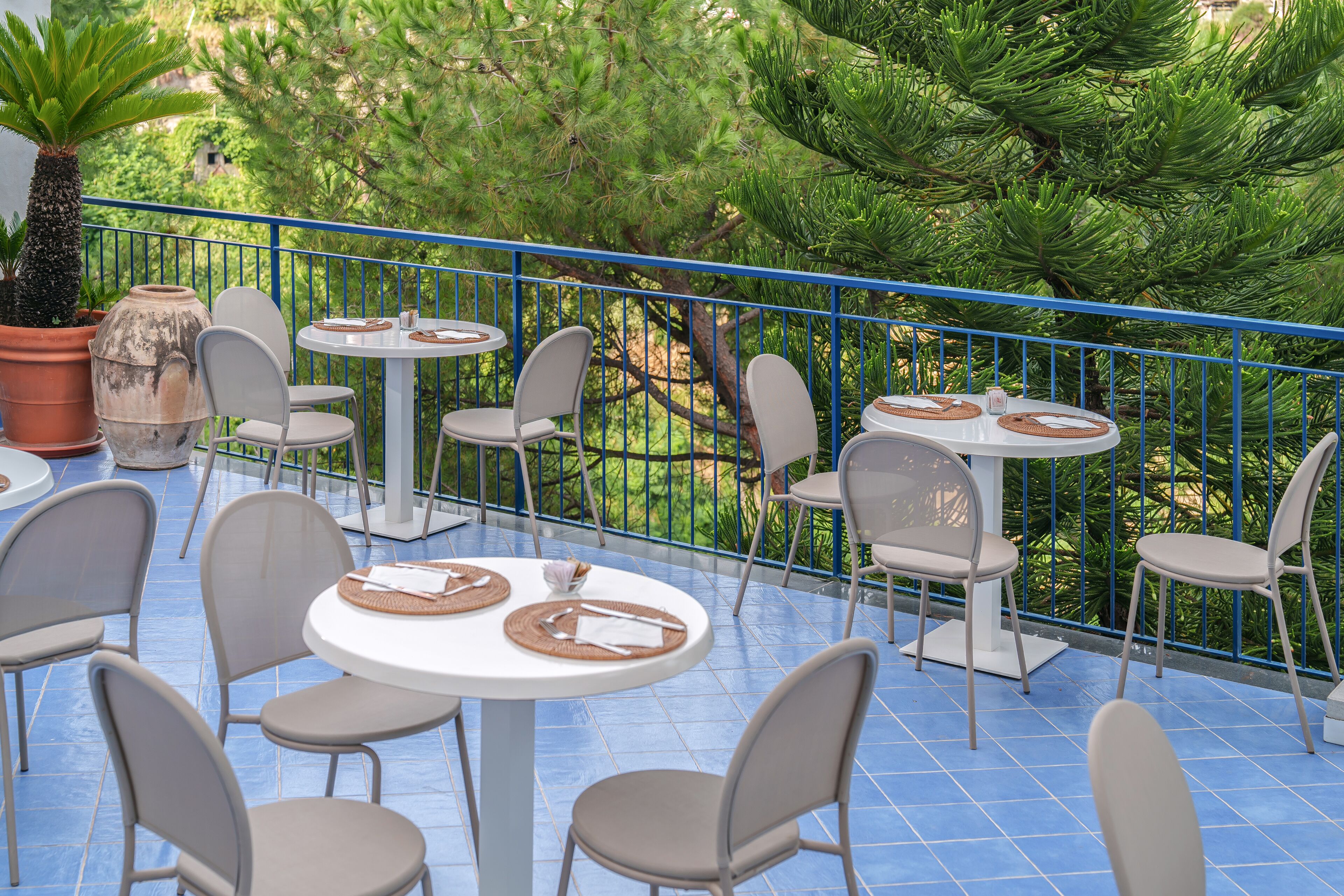 An outdoor patio with round tables and chairs, surrounded by a blue railing and lush greenery.