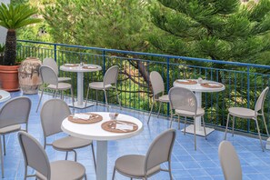 Un patio al aire libre con mesas y sillas redondas, rodeado por una barandilla azul y abundante vegetación.