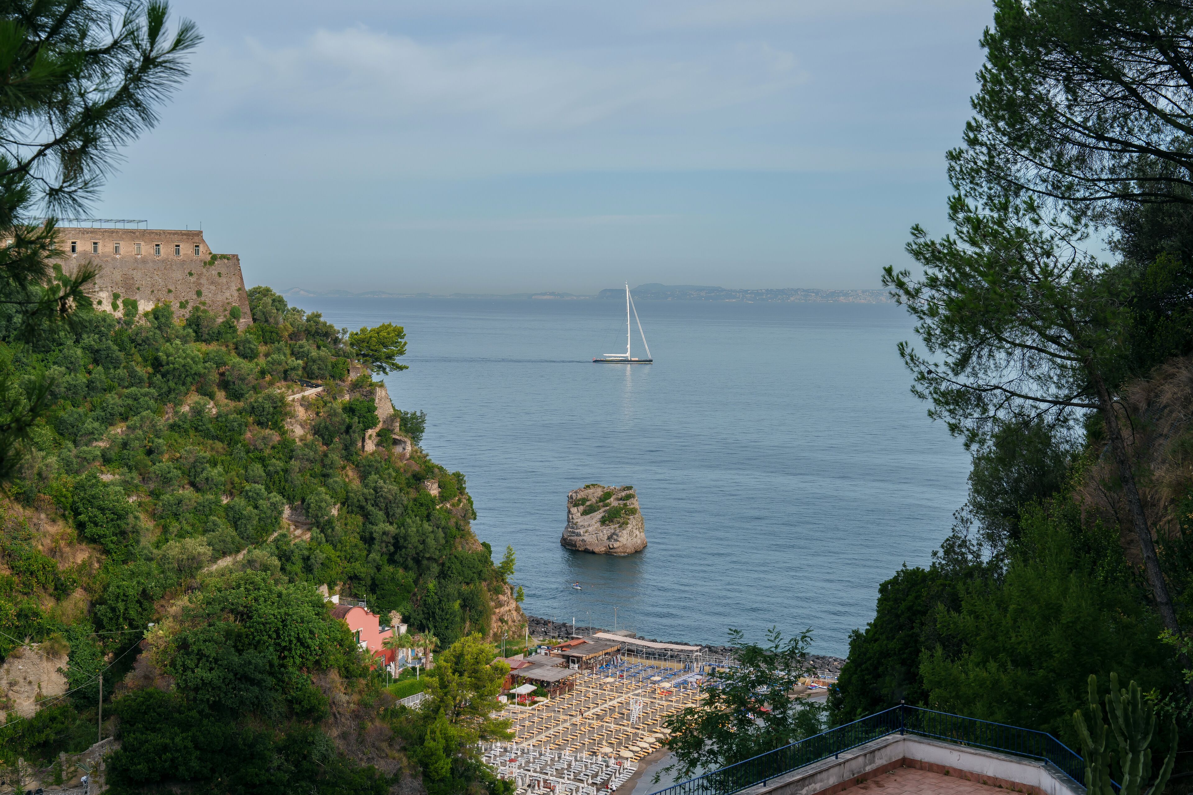 A coastal view with a castle, a sailboat, and a rocky outcrop.