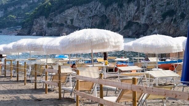 A beach with rows of sun loungers and thatched umbrellas, a rocky cliff in the background, and a calm sea. - Hotel Oriente (Vico Equense)