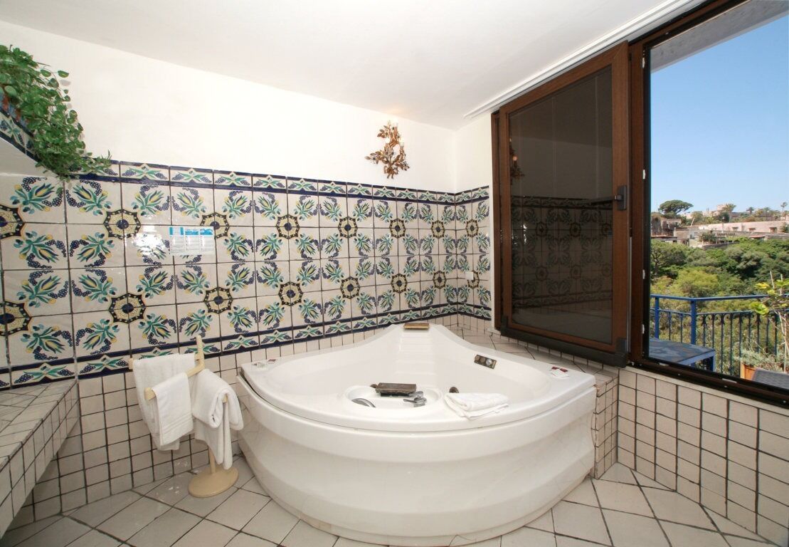 Un baño con bañera independiente, paredes de azulejos y vistas a través de un ventanal grande.