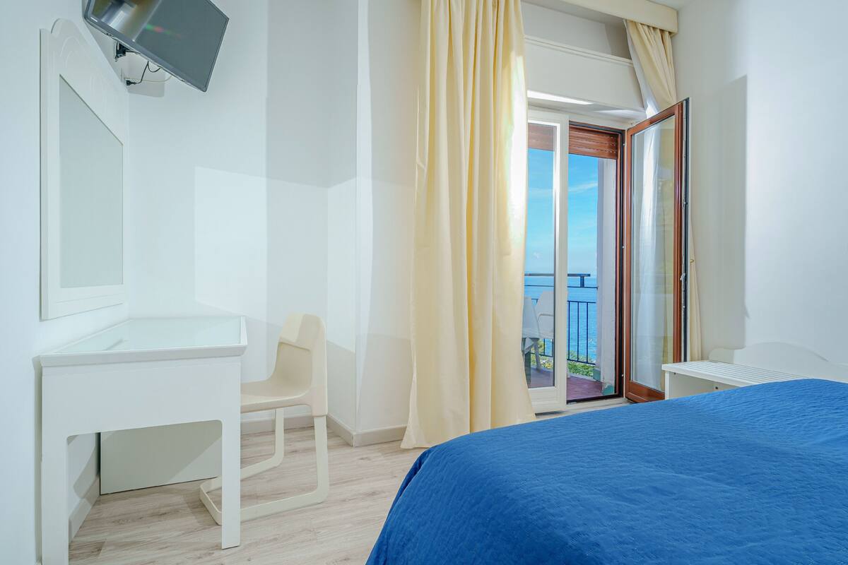 A hotel room with a bed, a desk, a chair, a TV, and a balcony with curtains.