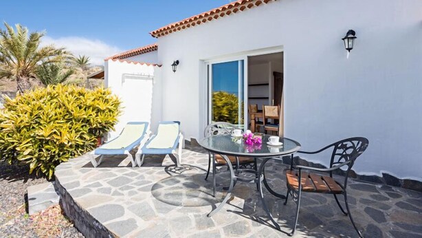 Un patio con una mesa y sillas, un edificio blanco con tejado de baldosas y una puerta que da al interior.