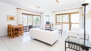 A spacious living area with a white sofa, a glass coffee table, and a dining table with chairs. There are large windows providing natural light and a view of greenery outside. - Residencial Las Norias (Los Llanos de Aridane)