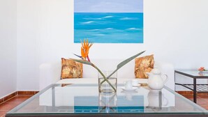 A modern room with a glass coffee table, a white sofa with patterned cushions, a painting of the ocean on the wall, and a small table with a teapot and cup. - Residencial Las Norias (Los Llanos de Aridane)
