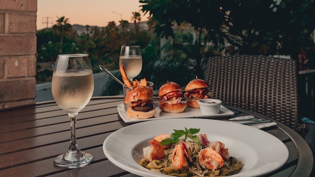 A dining table with a plate of seafood pasta, two burgers, a glass of white wine, and a glass of water. - Hyatt Place Los Cabos (San José del Cabo)