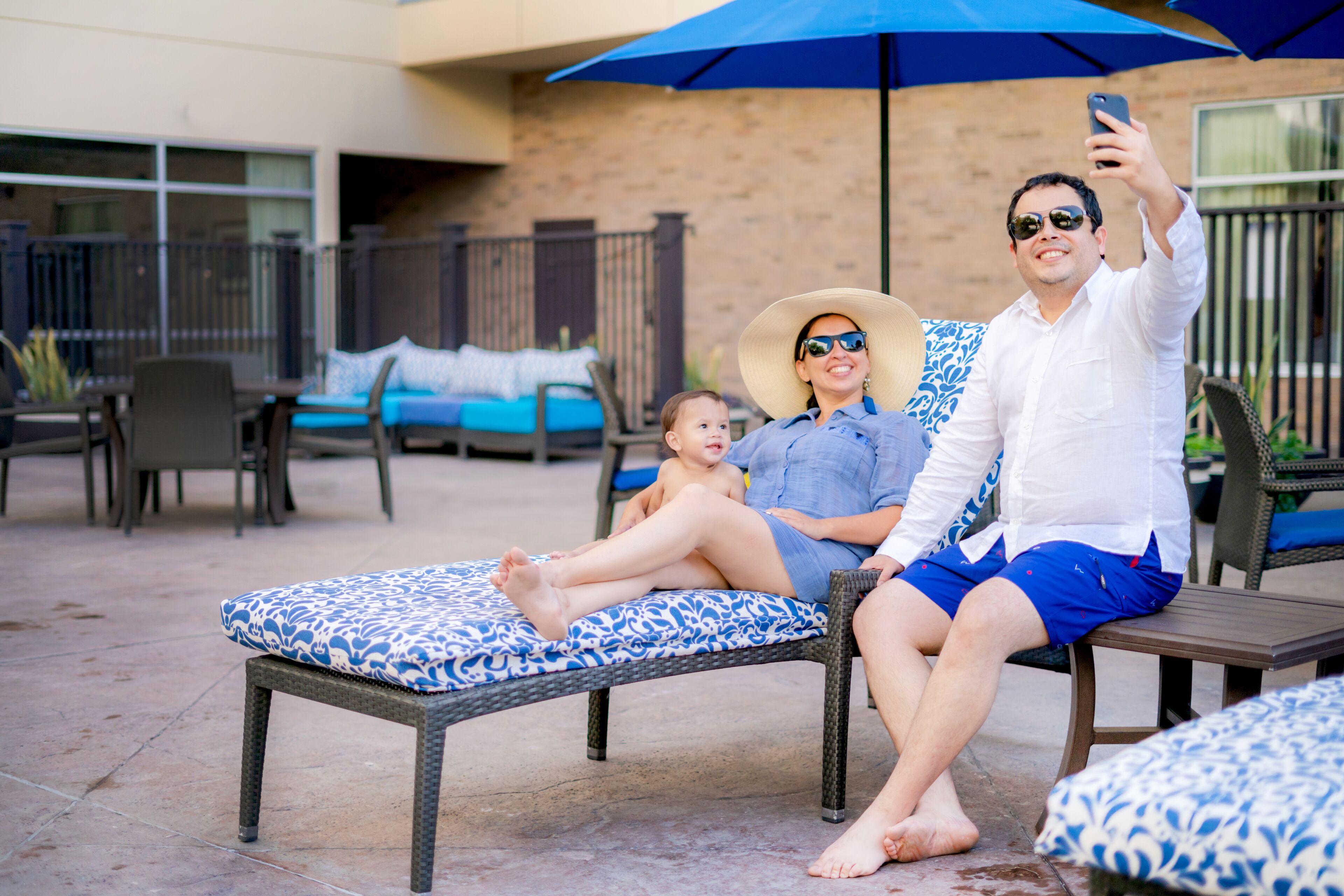 A couple relaxing on a wicker lounge chair with a blue and white patterned cushion, under a blue umbrella, in an outdoor patio area with wicker furniture and a brick wall in the background.