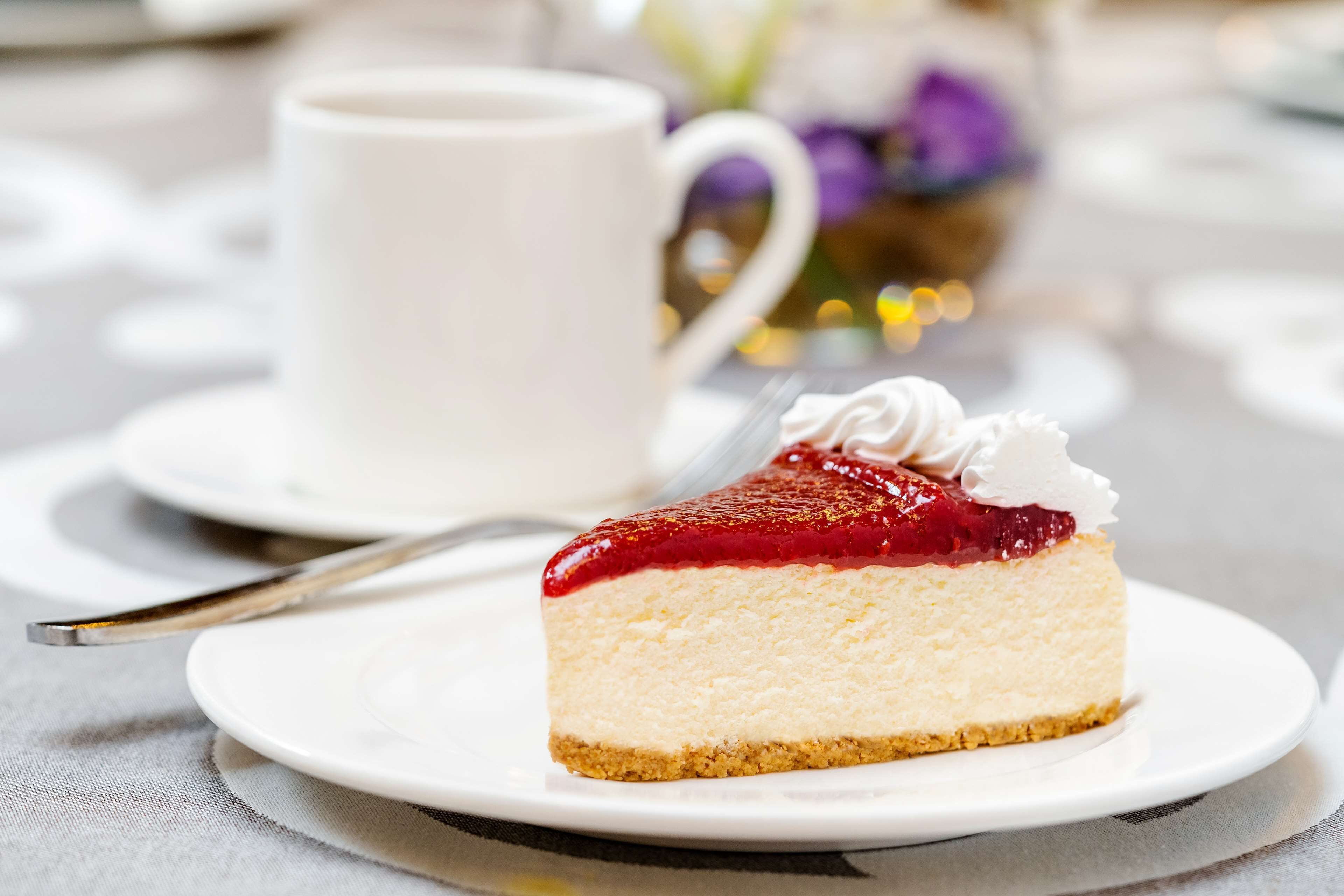 A slice of cheesecake with a red fruit topping and whipped cream, served on a white plate with a fork beside it.