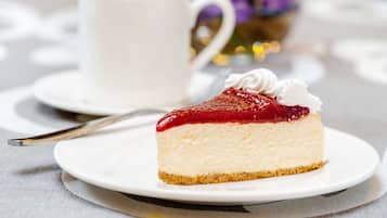 Ein Stück Käsekuchen mit roter Fruchtsoße und Schlagsahne, serviert auf einem weißen Teller mit einer Gabel daneben.