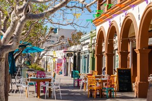 An outdoor café with tables and chairs, a tree, and buildings with arches and signs in the background. - Hyatt Place Los Cabos  (San José del Cabo)