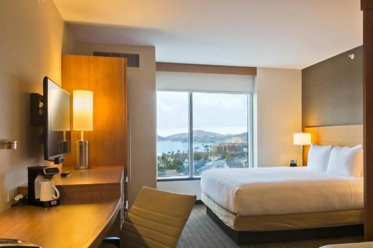 A hotel room with a bed, a desk with a lamp, a TV, and a view of the city.