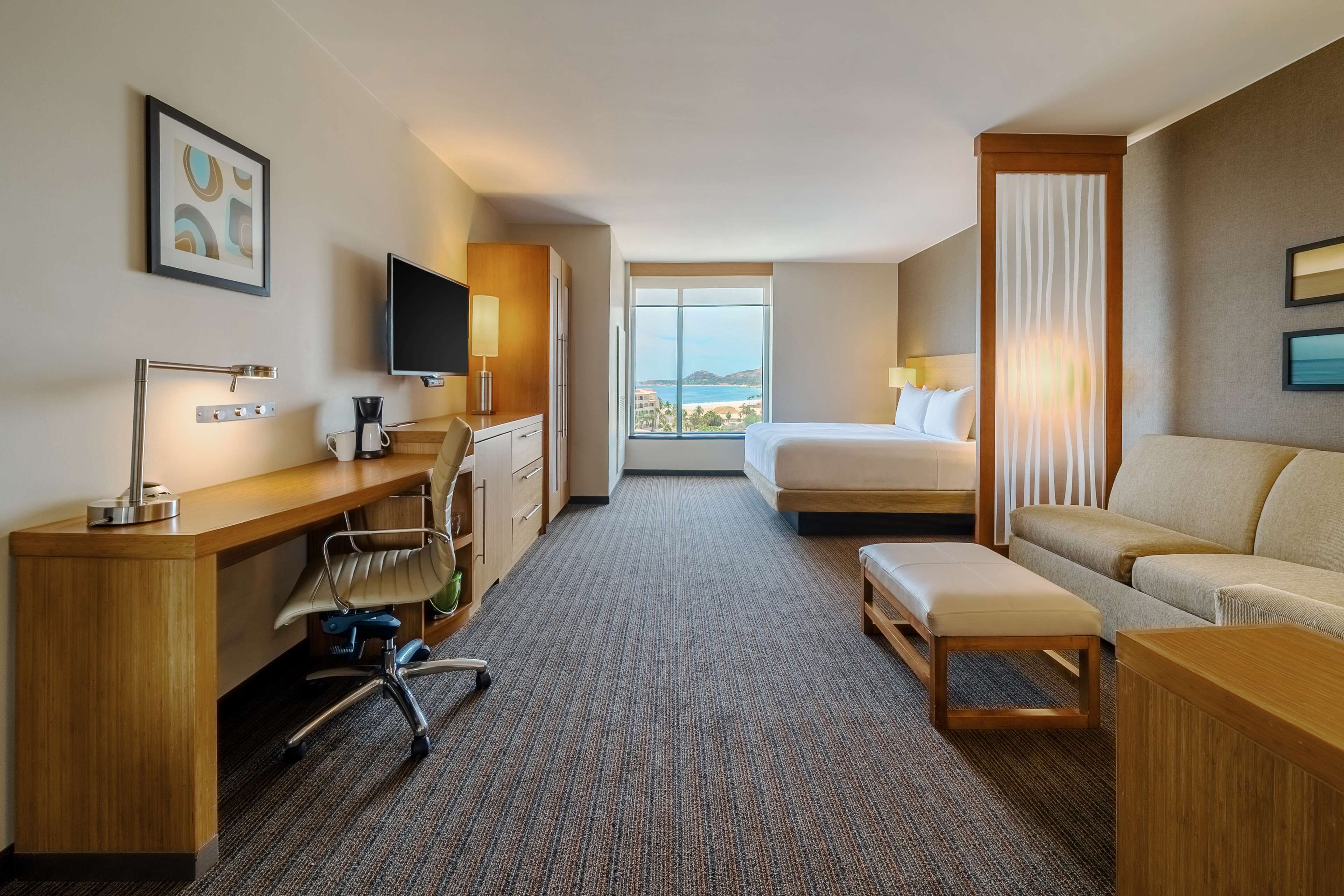A hotel room with a bed, a desk with a chair, a sofa, a TV, and a large window with a view.