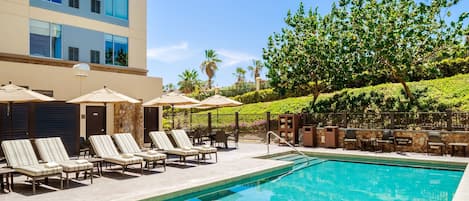 Una piscina con lettini e ombrelloni, circondata da un'area verde alberata e un edificio sullo sfondo.