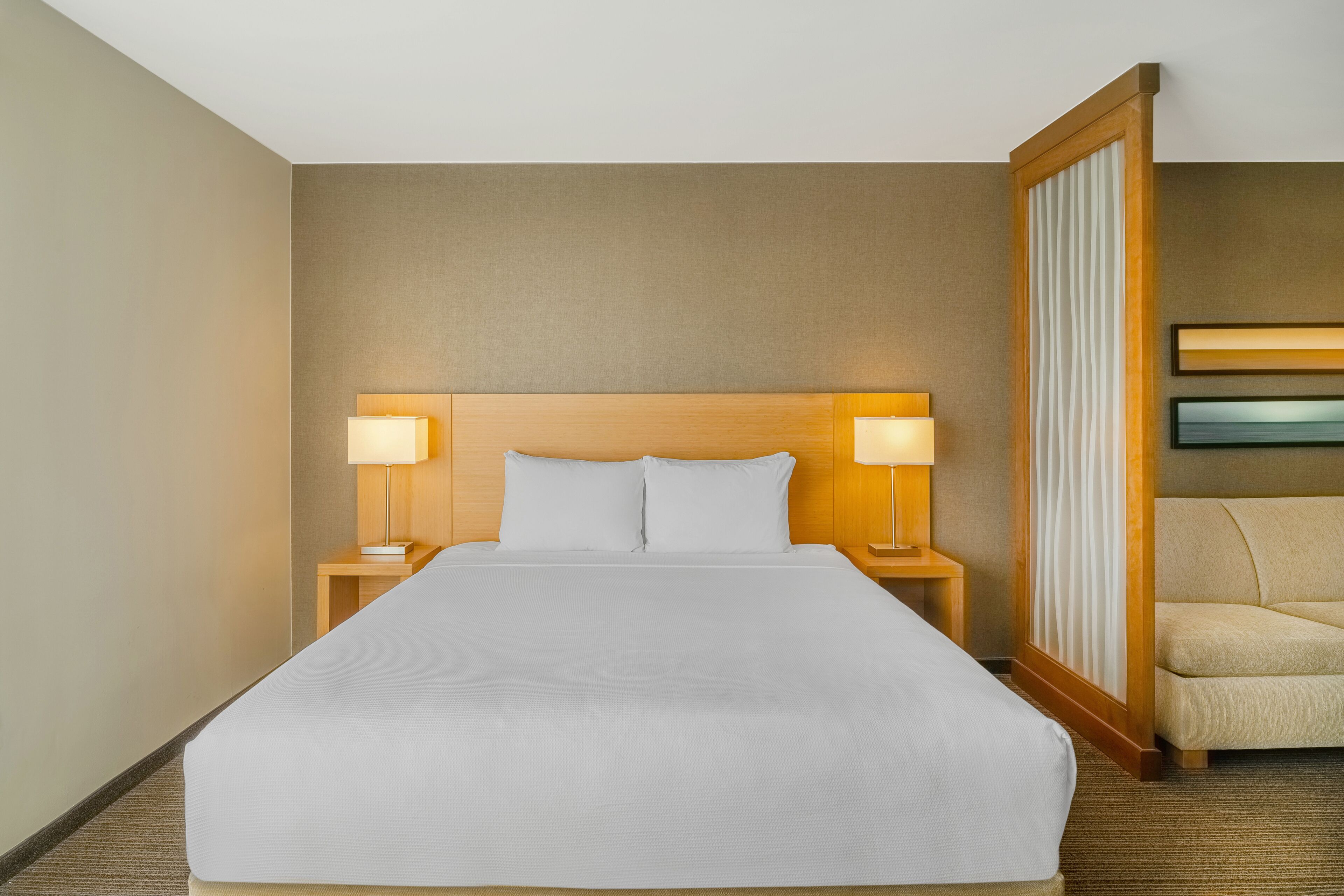 A hotel room with a large bed, two bedside lamps, a wooden headboard, a nightstand, a chair, and a wall-mounted TV.