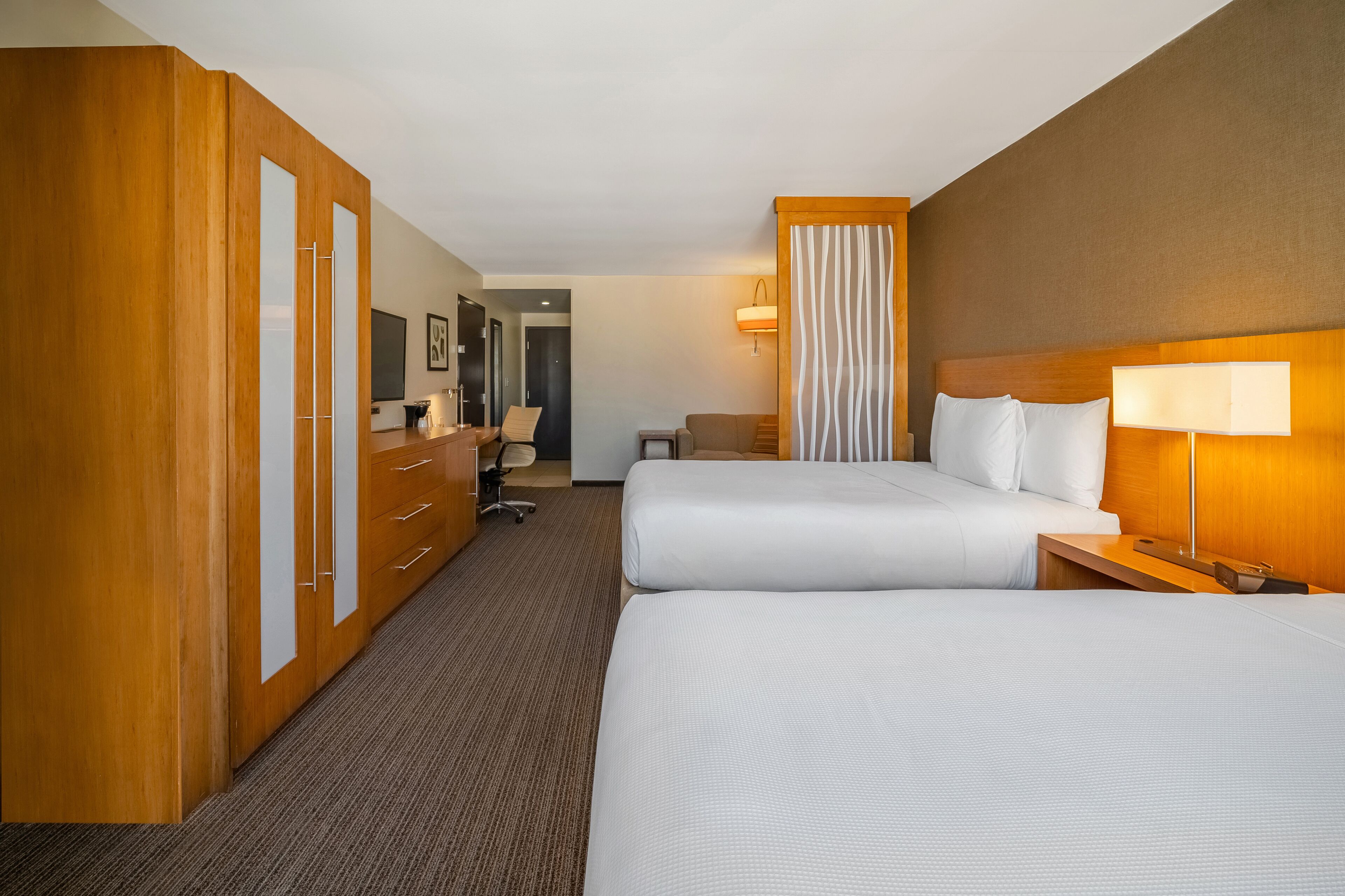 Habitación de hotel con dos camas, armario de madera, escritorio, silla, lámpara y vista al pasillo.