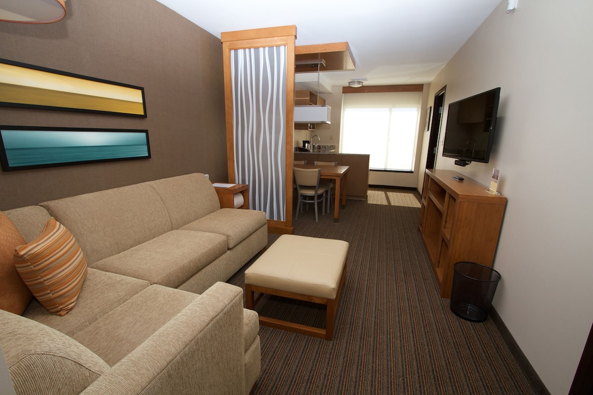 A compact hotel room with a sofa, a small table, a television, and a kitchenette area.