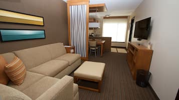 A compact hotel room with a sofa, a small table, a television, and a kitchenette area.