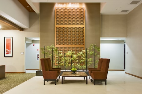 A lobby with a reception desk, two armchairs, a coffee table, a decorative wall panel, and a plant arrangement.