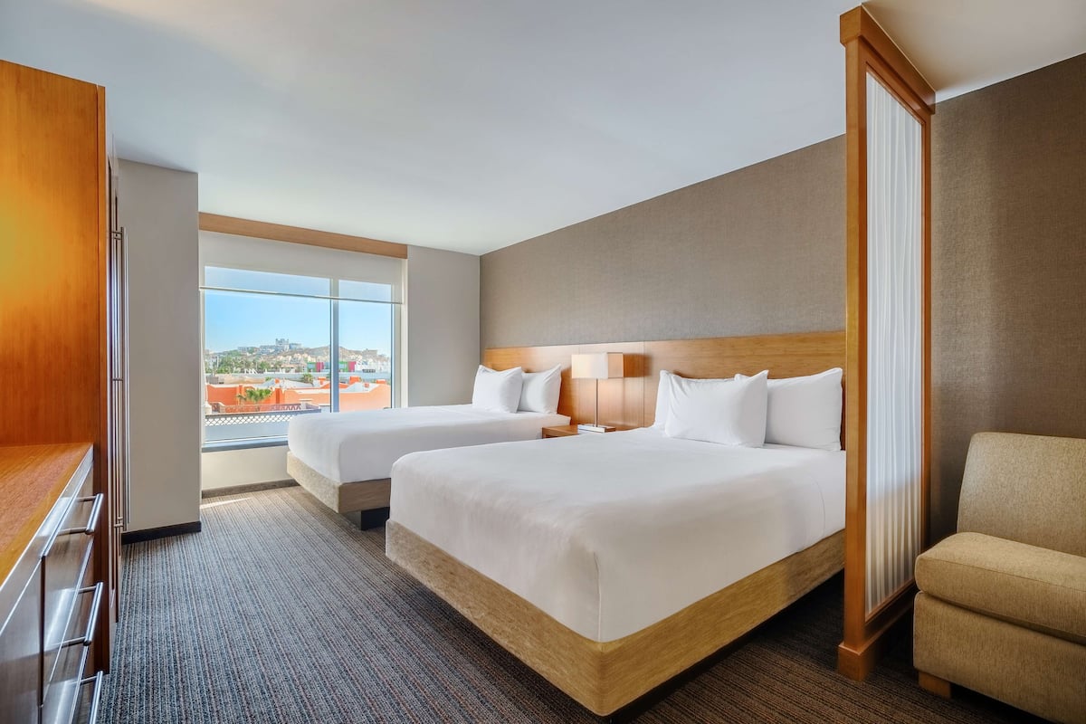 A hotel room with two beds, a wooden headboard, a large window with a city view, and a seating area with a chair.
