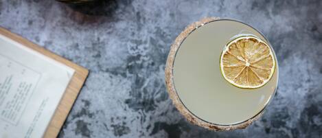 A top-down view of a cocktail with a slice of dried lemon on the rim, a bowl of potato chips, and a menu on a table.