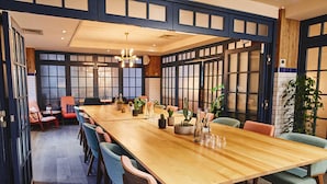 A long wooden dining table with chairs, surrounded by large windows and potted plants. - The Corner London City (London)