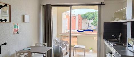 A compact room with a sliding glass door leading to a balcony, a small dining table, and a kitchenette area.