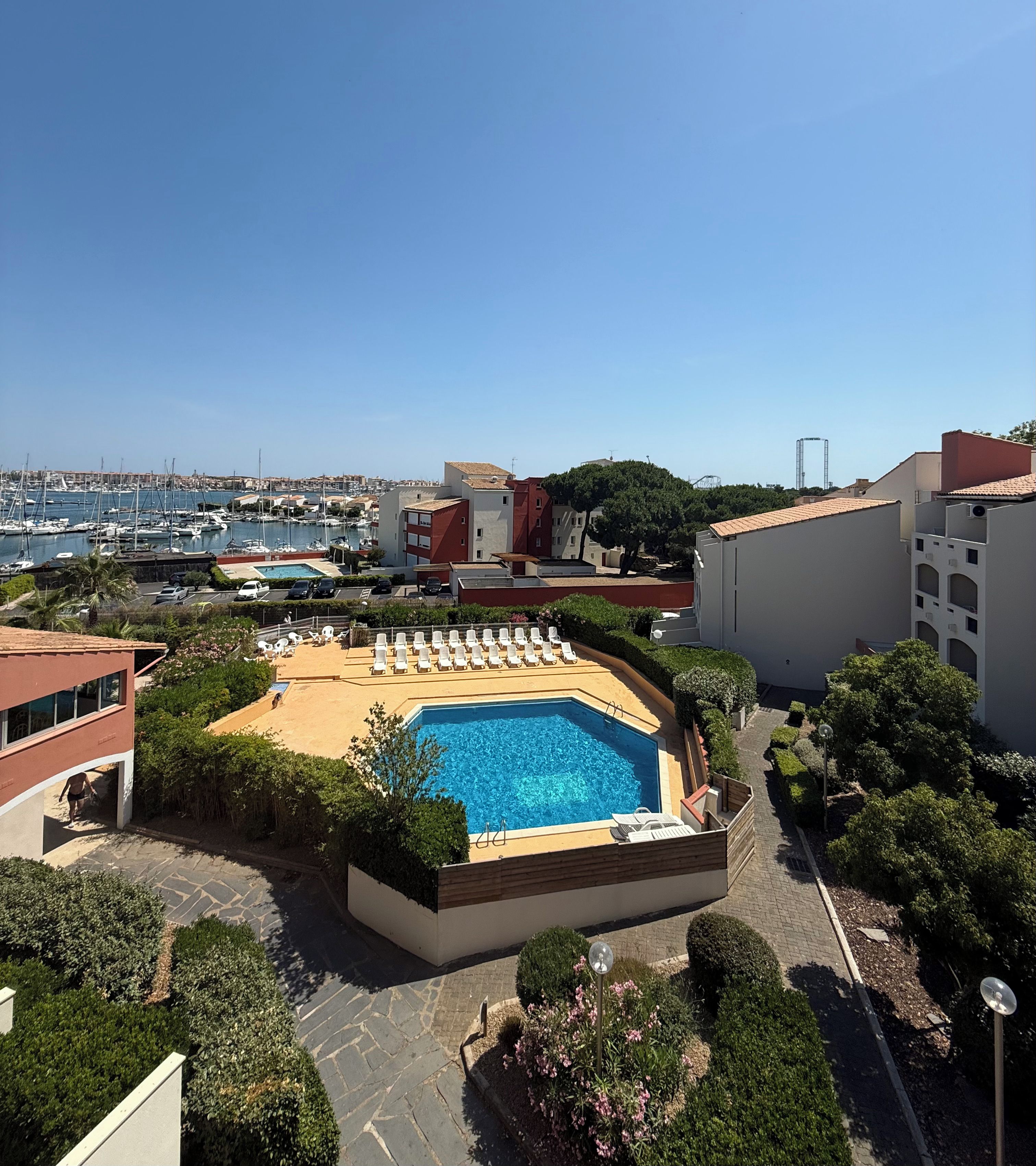 Una piscina con tumbonas, rodeada de jardines con vistas a un puerto deportivo y edificios al fondo.