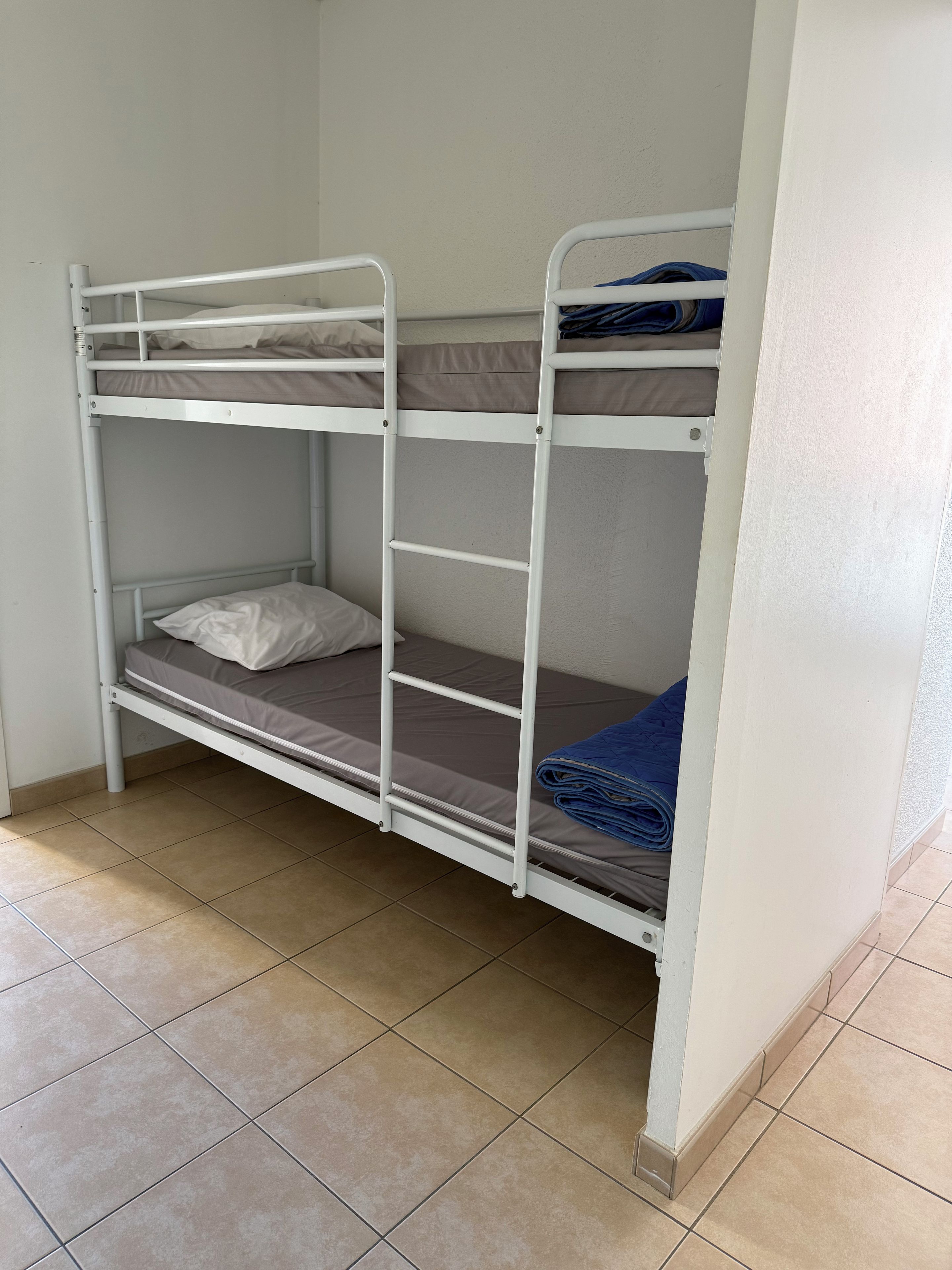 A bunk bed with two levels, each with a mattress and pillows, in a room with tiled flooring.