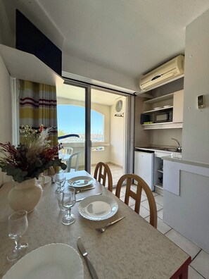 Un coin repas avec une table dressée pour quatre personnes, une cuisine équipée d’un four à micro-ondes et d’un évier, et une vue sur la mer depuis la porte-fenêtre donnant sur le balcon.