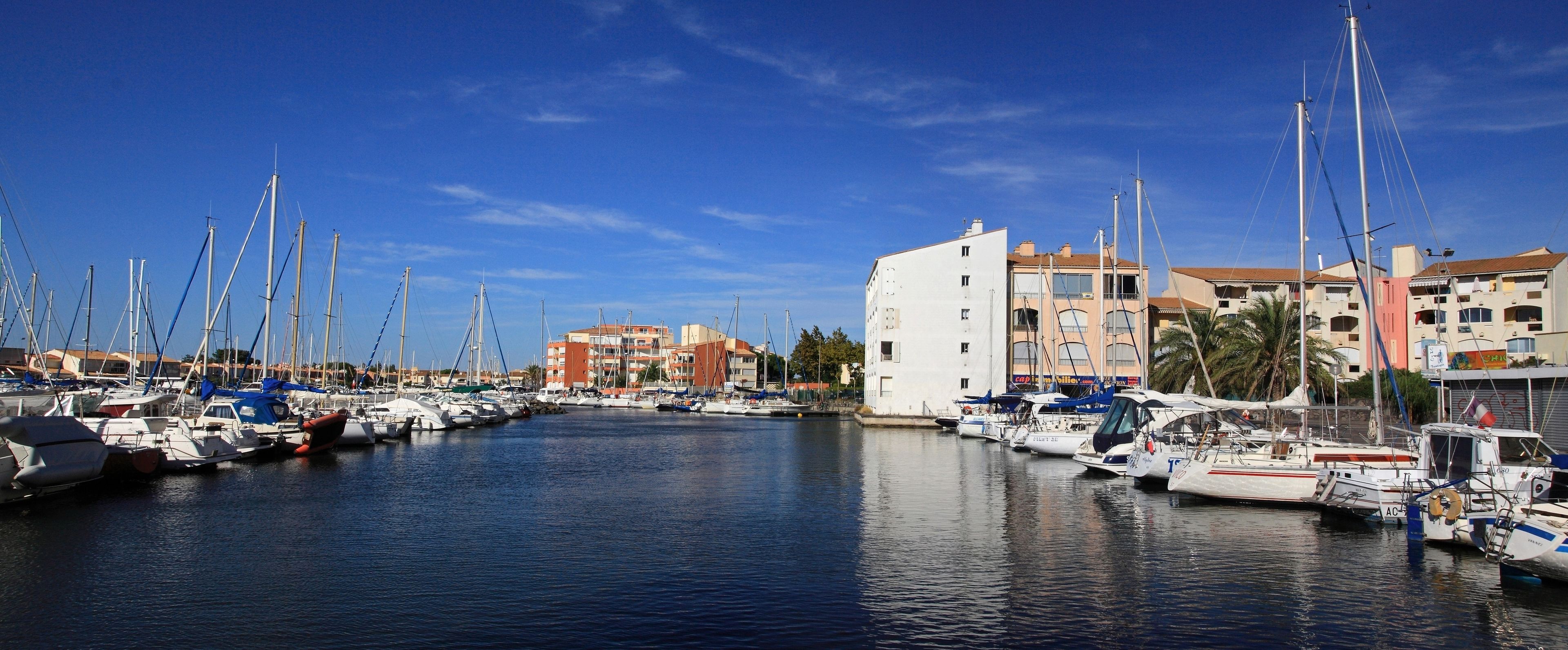Un puerto deportivo con varias embarcaciones amarradas, edificios al fondo y aguas tranquilas.