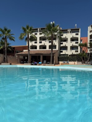 A swimming pool with a clear blue surface, surrounded by palm trees and a multi-story building with balconies. - Résidence Agathéa (Agde)