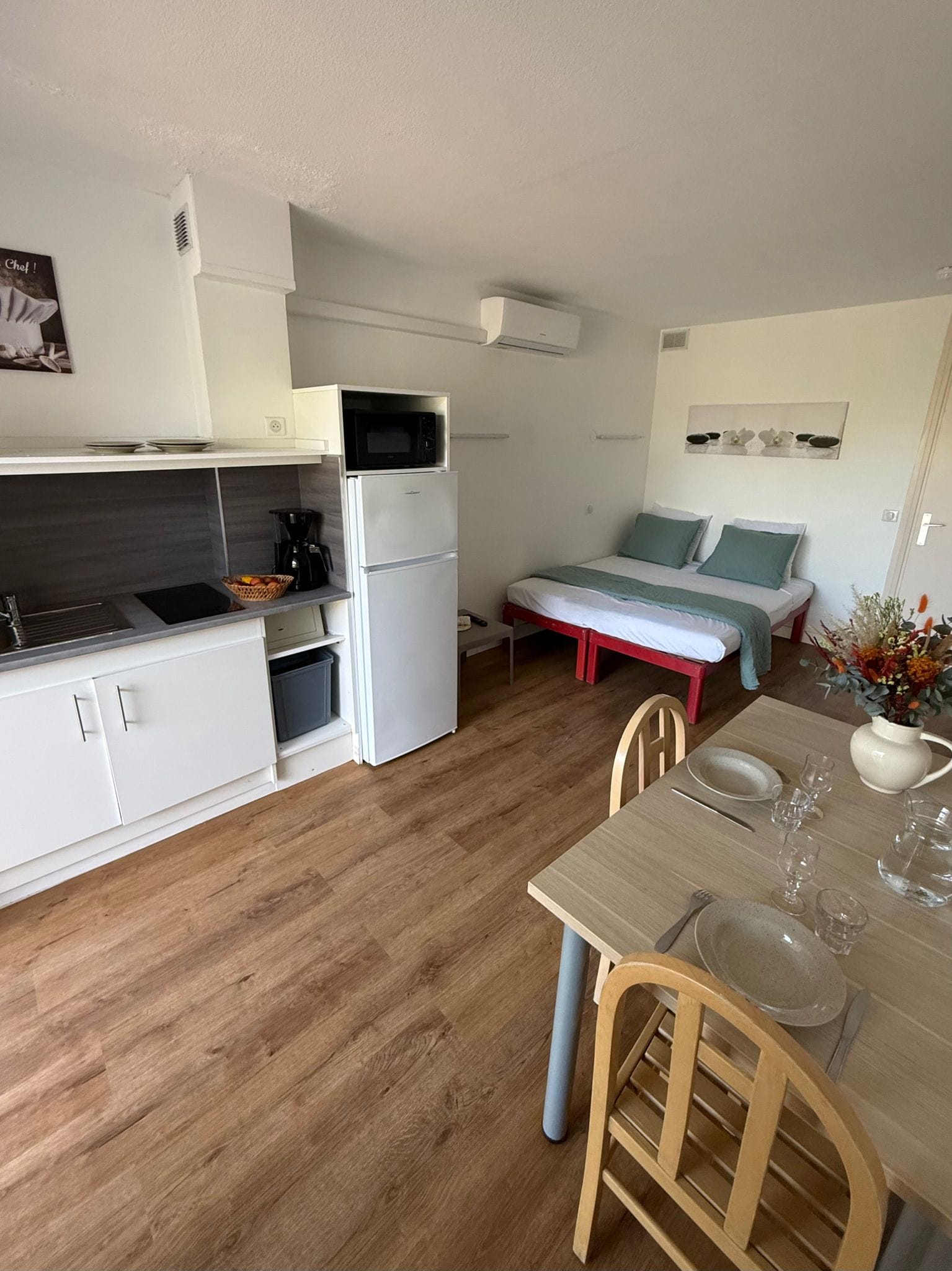 A compact kitchen with a microwave, coffee maker, and a small refrigerator. There is a dining area with a table, chairs, and a vase with flowers.