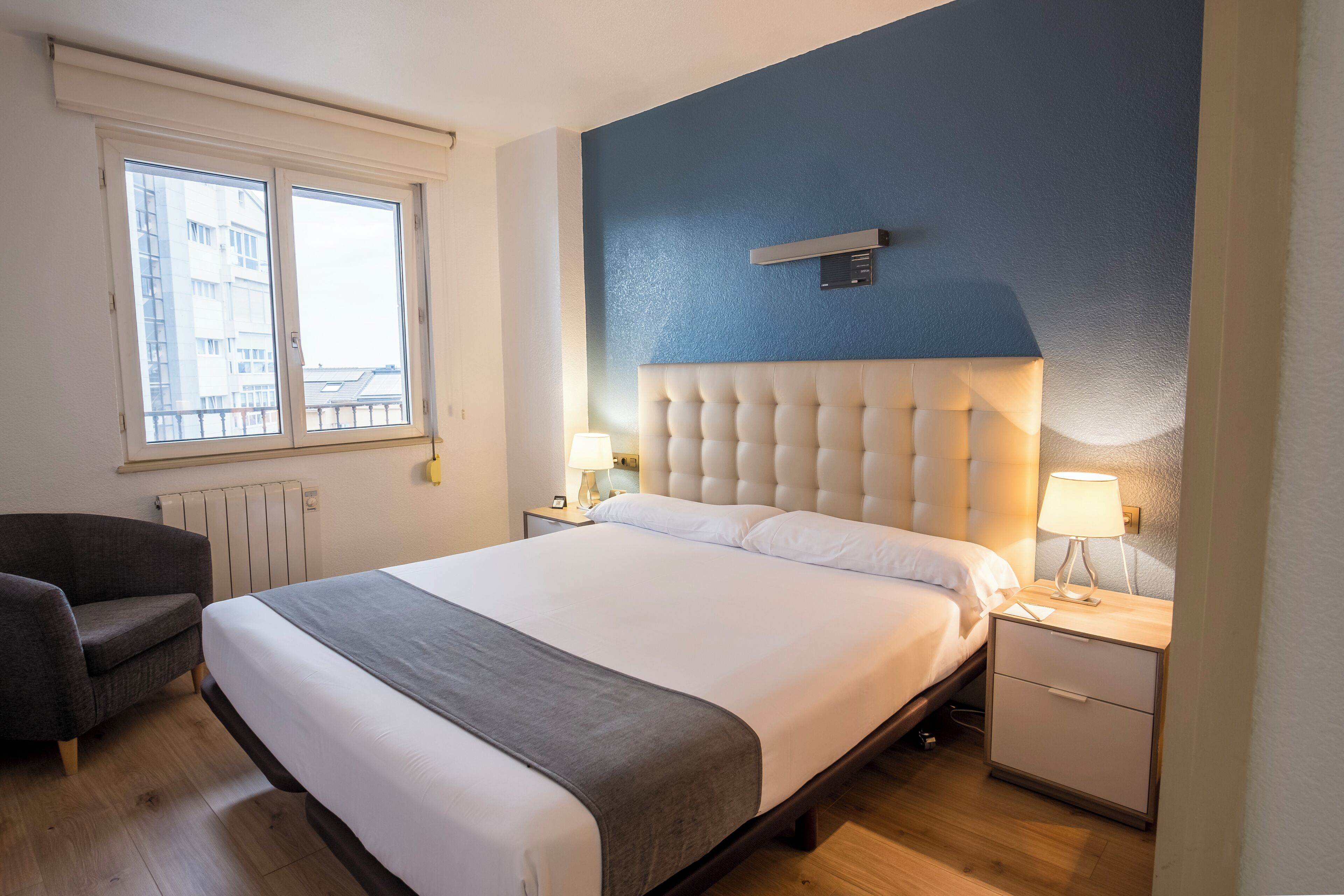 Una habitación de hotel con una cama grande, una mesita de noche con lámpara, una silla y una ventana con vistas a edificios.