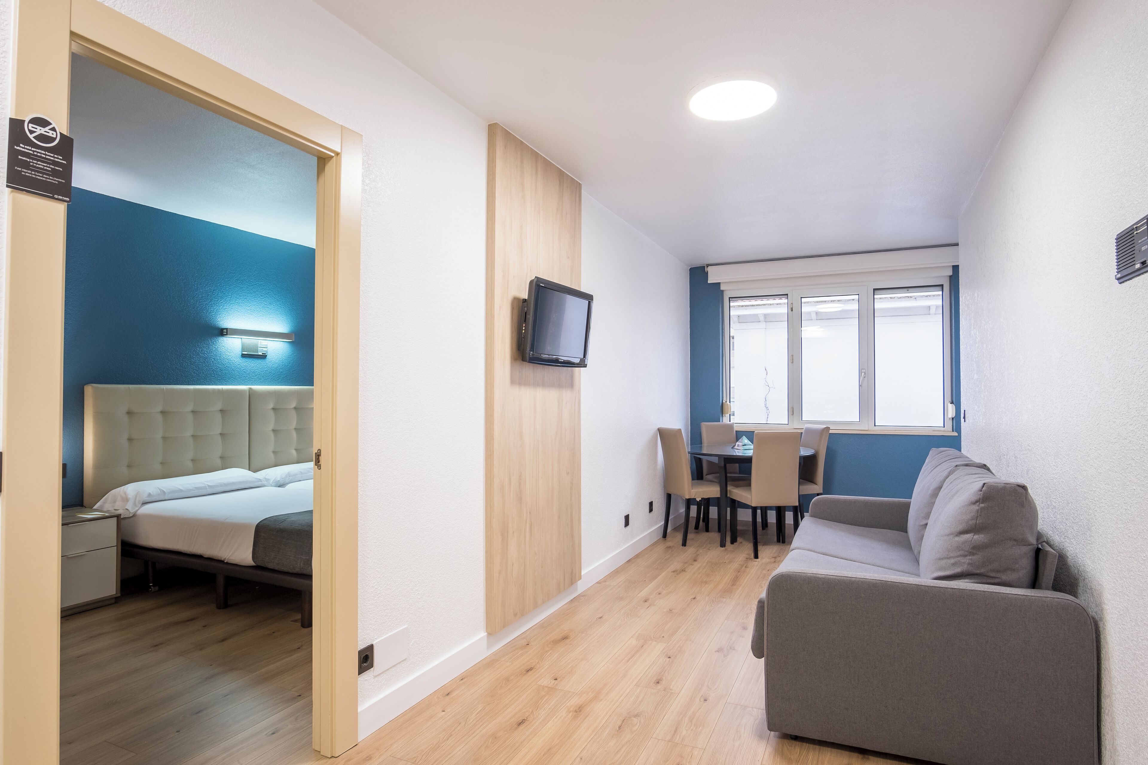Una habitación de hotel moderna con una cama, un televisor montado en la pared, un rincón de comedor con mesa y sillas, y un sofá.