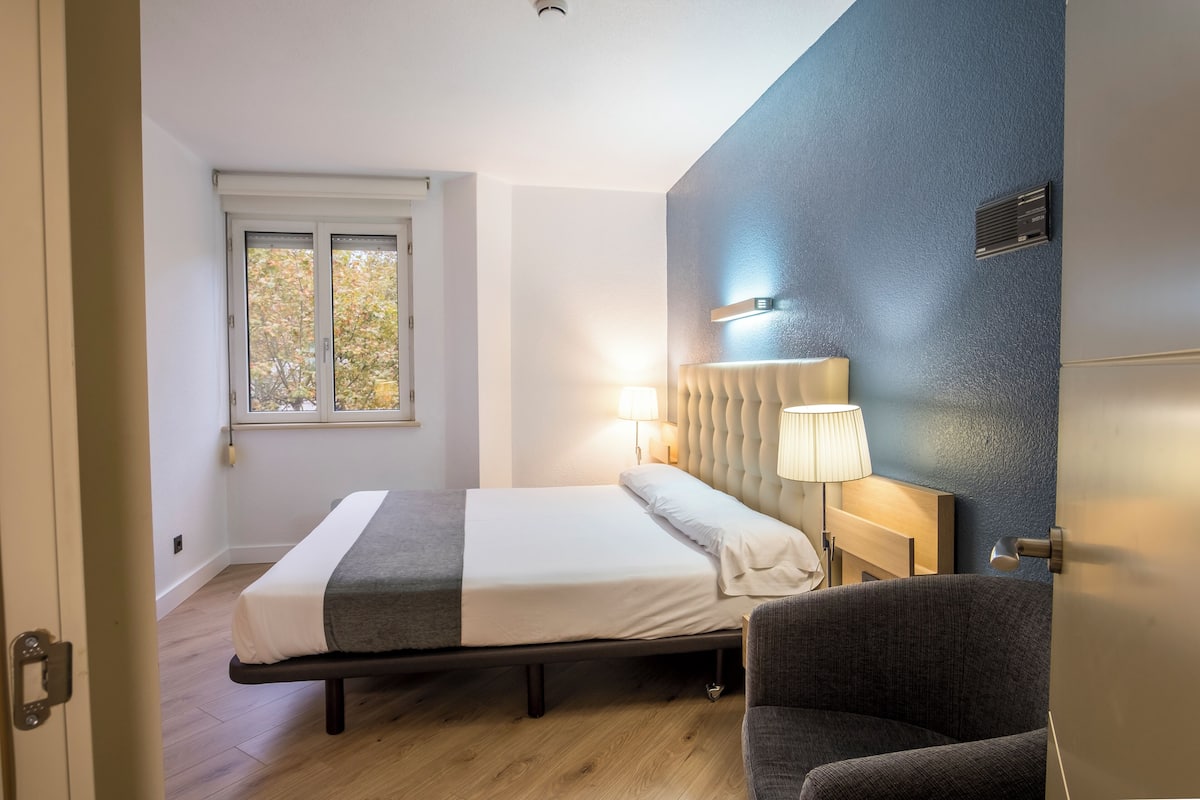 A modern hotel room with a bed, a chair, a window, and a wall-mounted lamp.