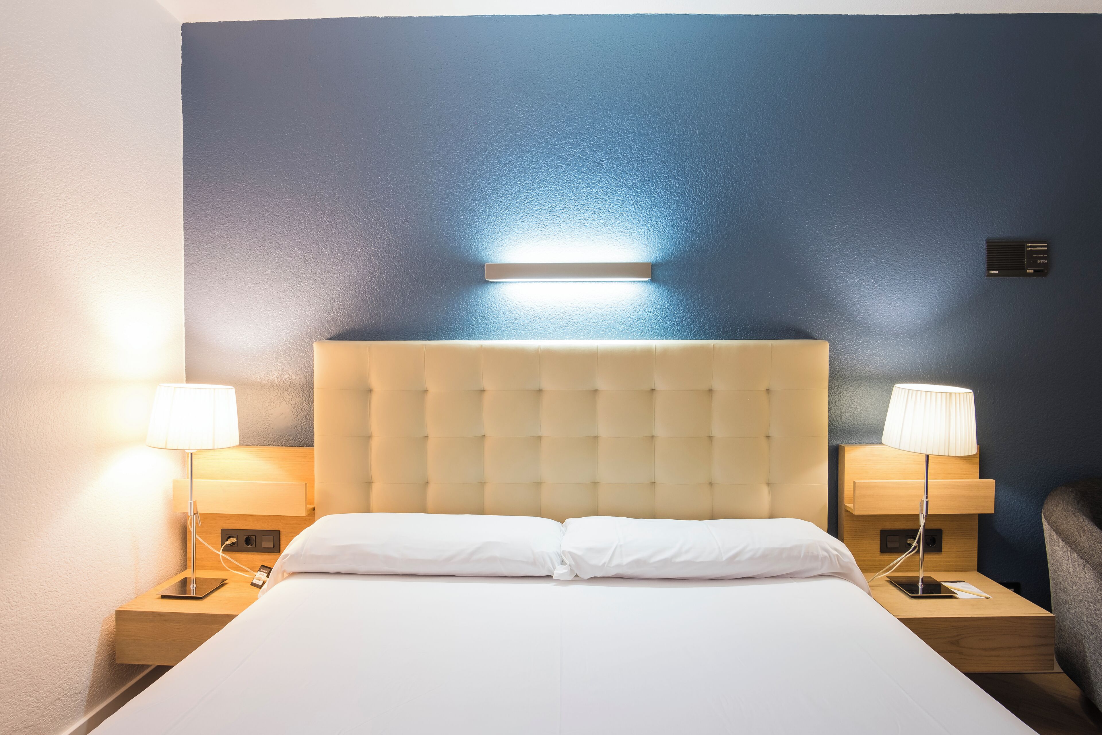 Una habitación de hotel con una cama grande, dos mesitas de noche con lámparas, una luz empotrada en la pared sobre la cama y un equipo de aire acondicionado de pequeño tamaño en la pared.