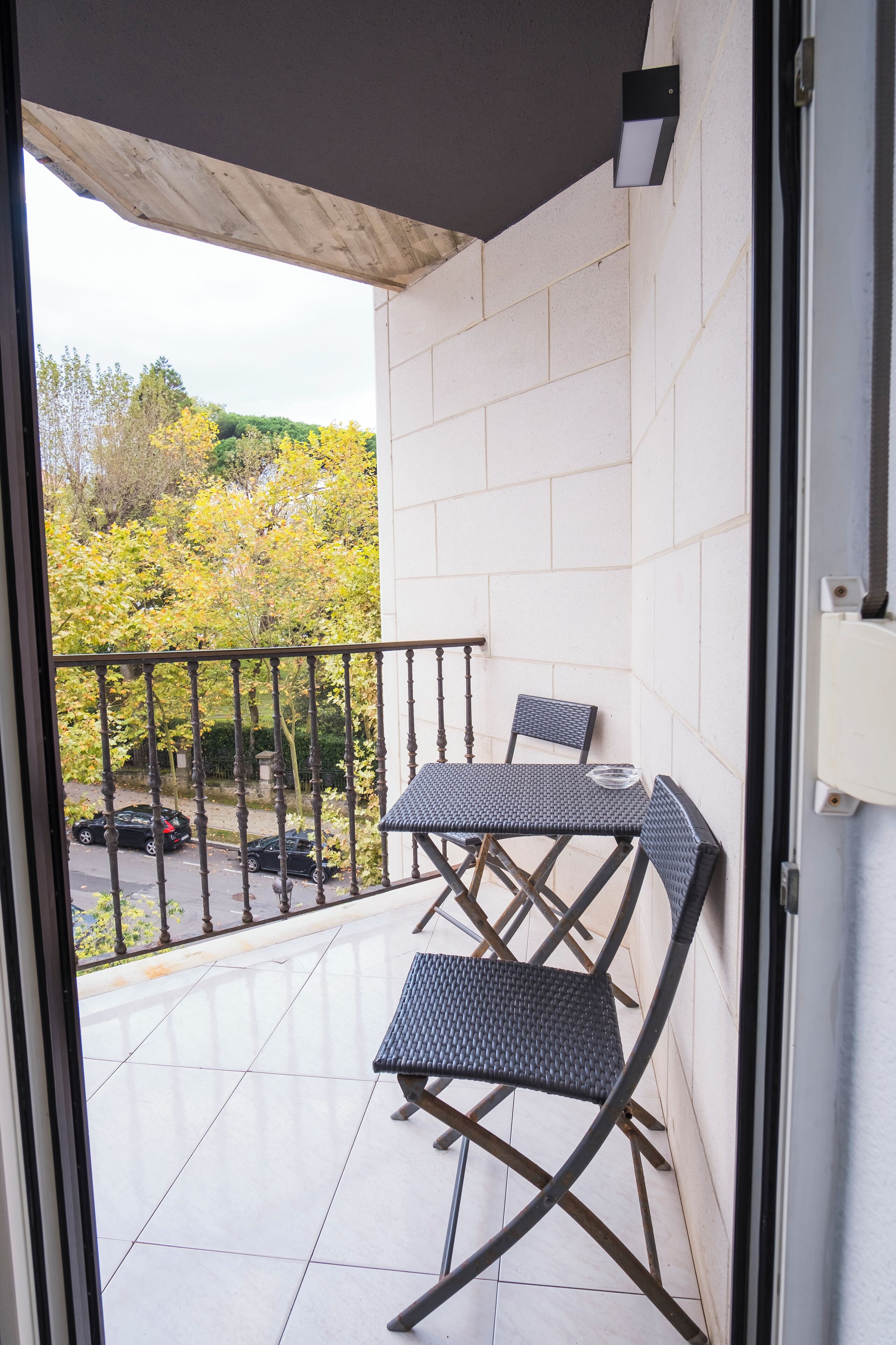 A balcony with a table and chairs, a view of trees and parked cars, and a tiled floor.