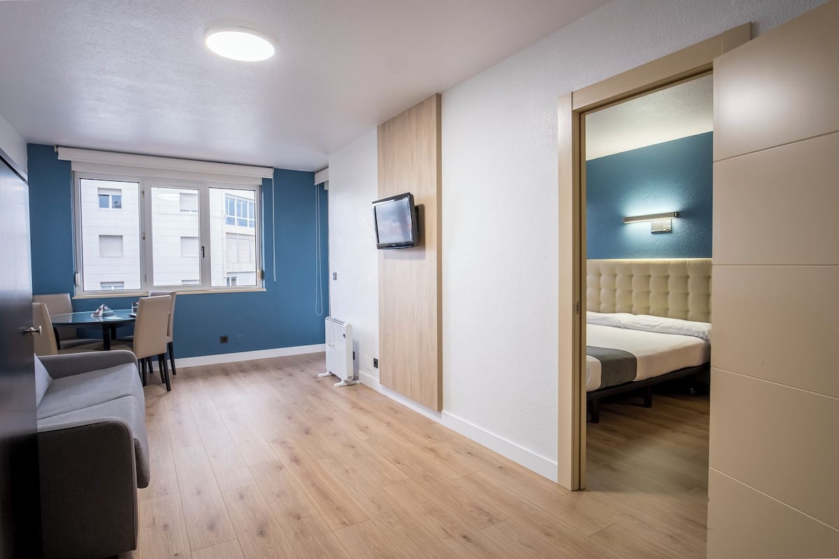 A modern room with a bed, a TV mounted on the wall, a dining area with a table and chairs, and a view of the outside through a window.
