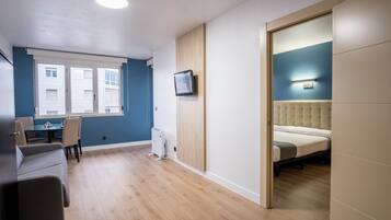 Habitación moderna con cama, televisor montado en la pared, zona de comedor con mesa y sillas, y vista al exterior a través de una ventana.