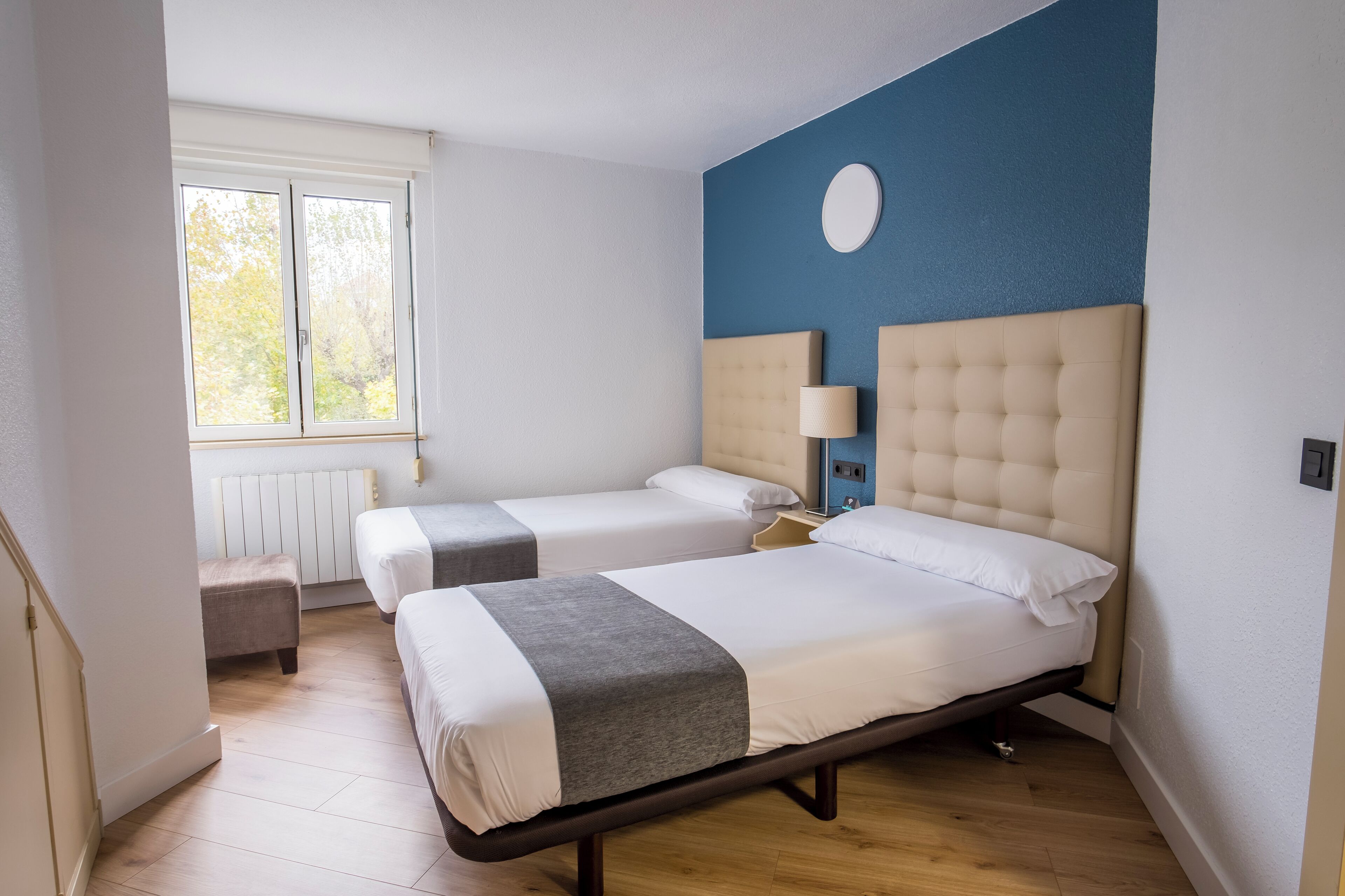A hotel room with two beds, a window, a radiator, and a blue accent wall.