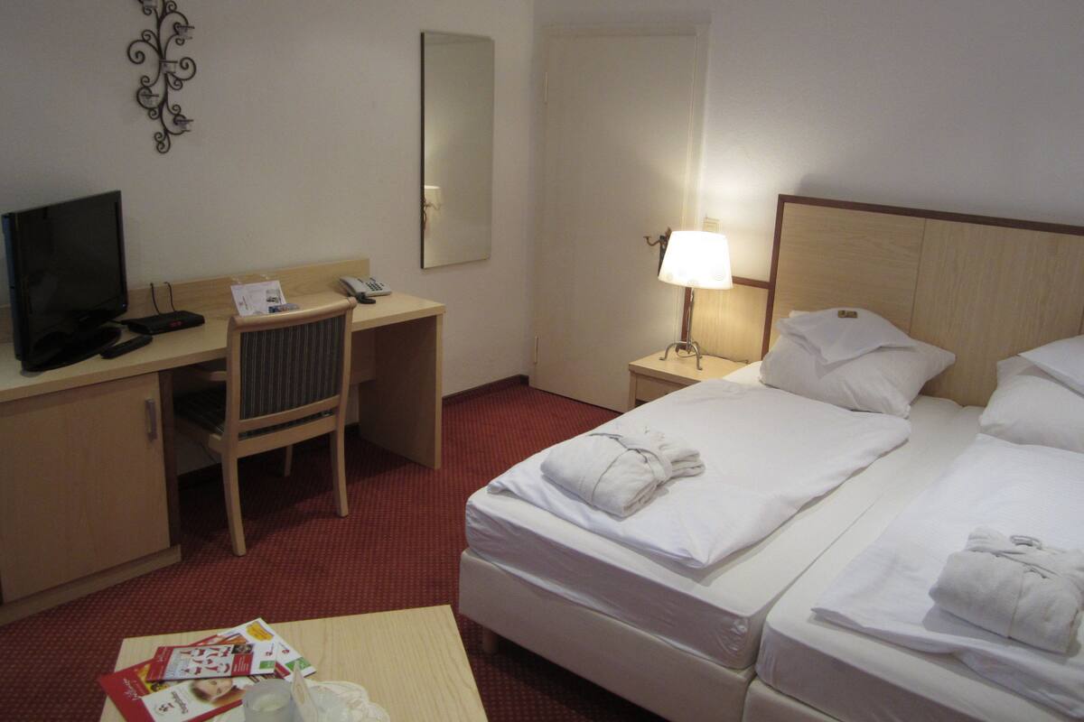 A hotel room with a bed, a desk with a computer, a chair, a TV, and a bedside table with a lamp.