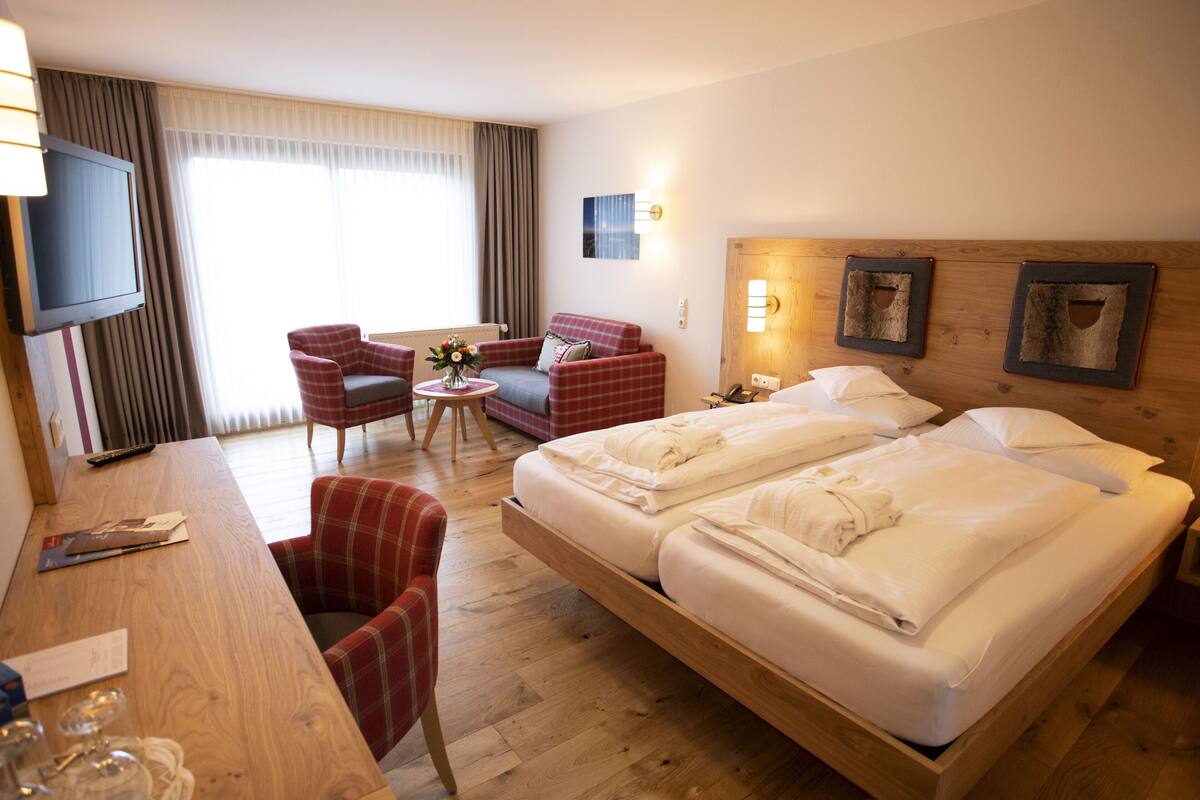 A hotel room with a large bed, a desk with a TV, a red armchair, and a small table with a vase.