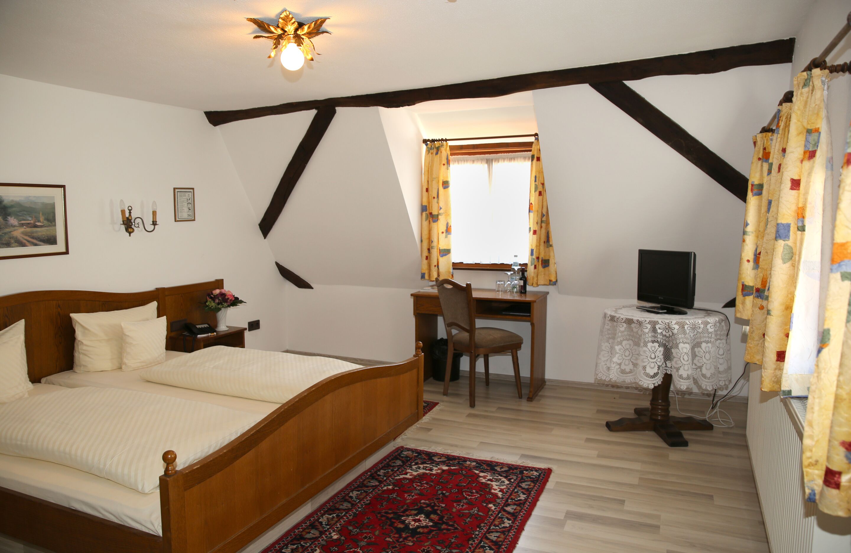 Une chambre à coucher avec un lit en bois, un bureau, une chaise, une petite table sur laquelle se trouve un téléviseur, et une fenêtre avec des rideaux.