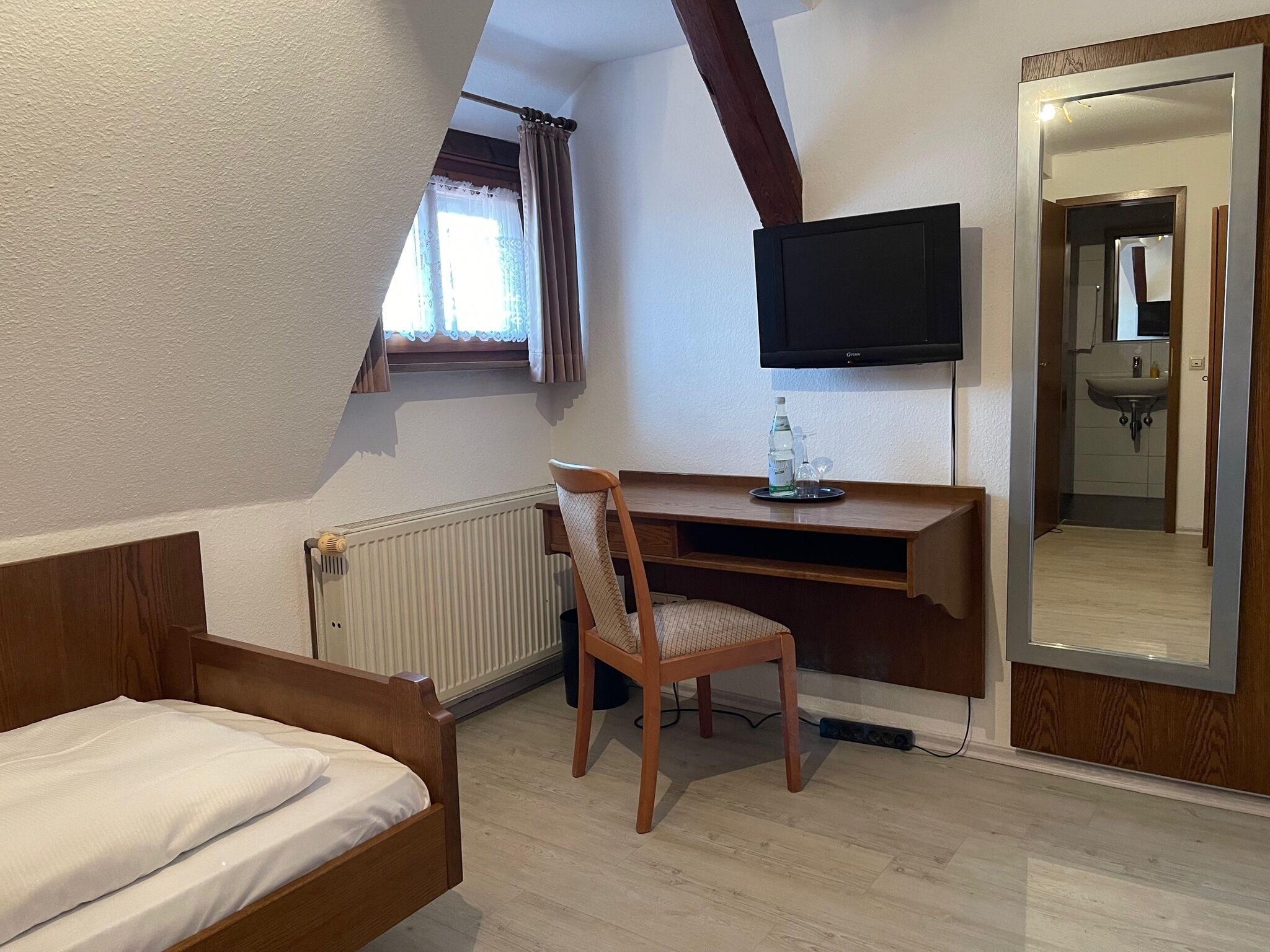 A bedroom with a bed, a desk with a chair, a television, and a bathroom visible through a mirror.