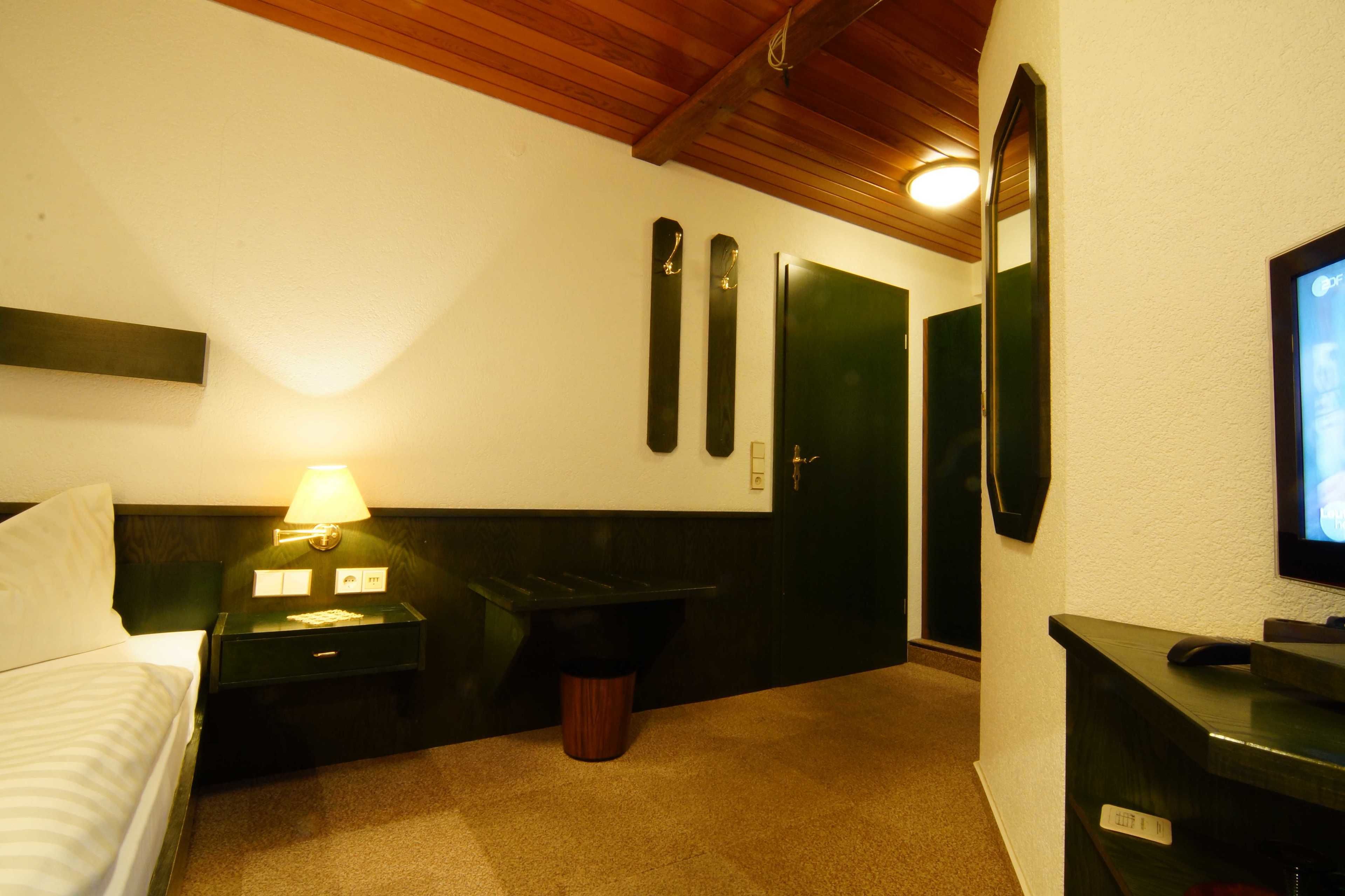 Una habitación de hotel con una cama, una mesita de noche con lámpara, un cubo de basura, una puerta negra y un televisor sobre un mueble.