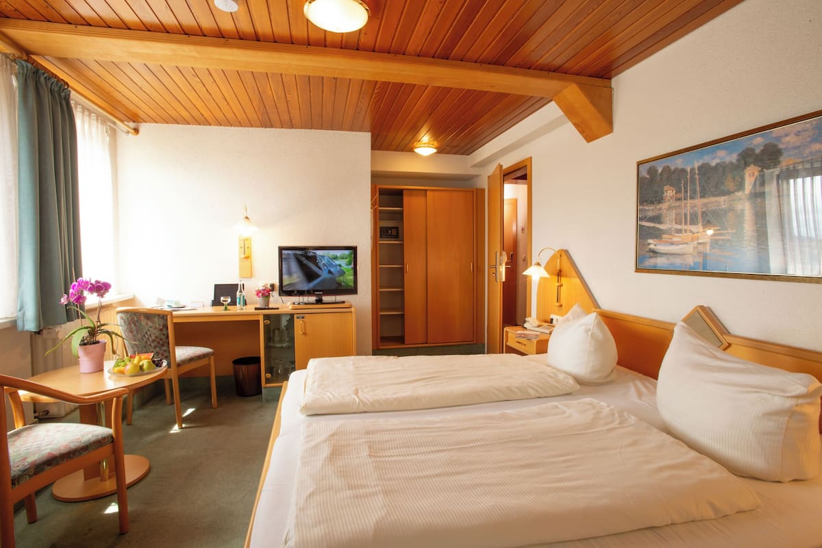 A hotel room with a wooden ceiling, a bed with white linens, a desk with a computer and a TV, a chair, a small table with a plant, and a painting on the wall.
